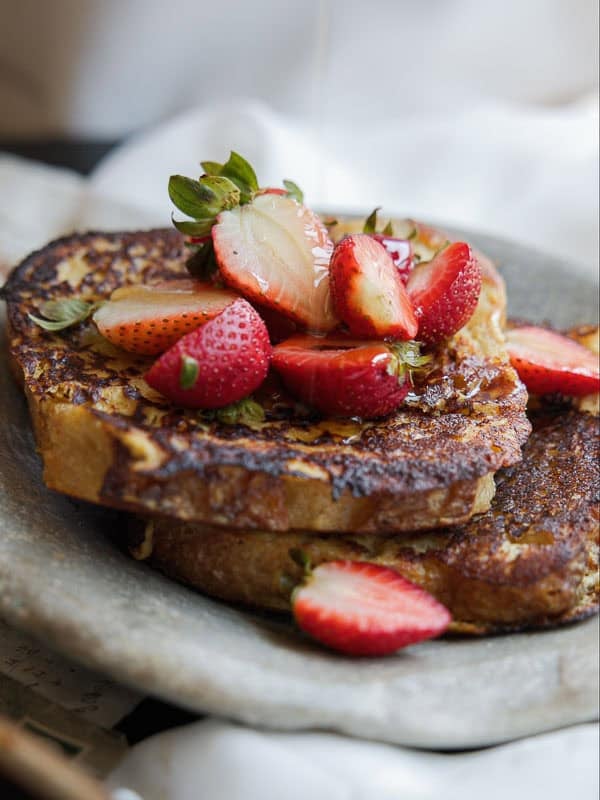 Two thick slices of vegan French toast topped with fresh, sliced strawberries and a drizzle of syrup, served on a rustic stone plate.